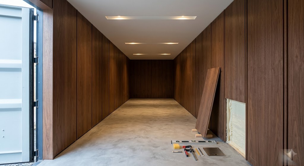 Pod interior near end of fit-out: walnut walls and off-white ceiling almost complete, one panel still to install with finishing tools laid out on microcement floor